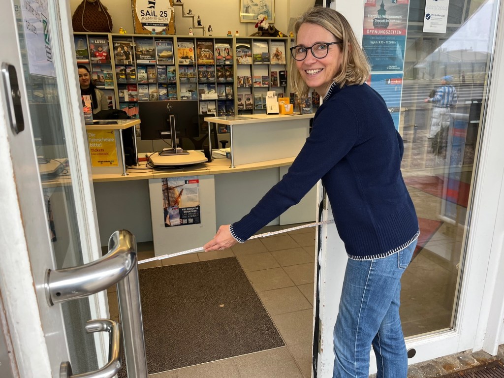 Eine Person steht vor einer Tür und misst die Breite mit einem Zollstock. Im Hintergrund ist ein Empfangstresen mit Informationsmaterialien zu sehen.