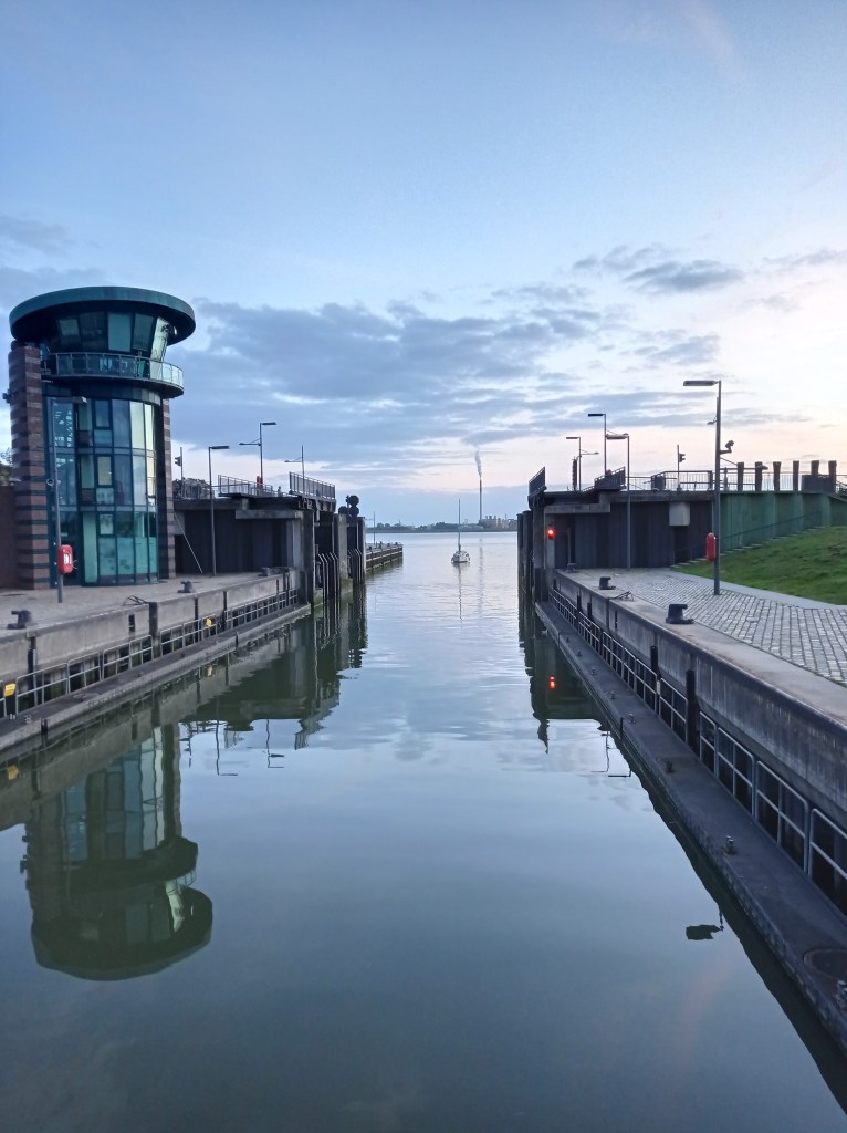 Blick auf die Schleuse am Neuen Hafen mit Kontrollturm und einem Schiff in der Schleusenkammer, bei Sonnenaufgang.