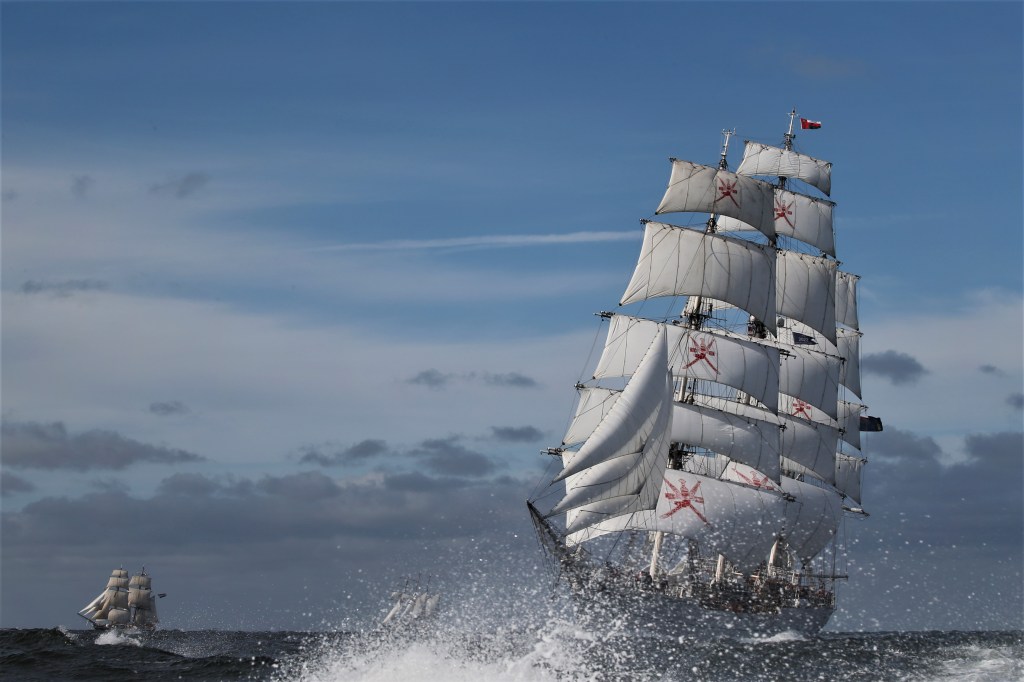 Ein großes Segelschiff fährt mit aufgeregten Wellen im Hintergrund auf dem offenen Meer unter einem bewölkten Himmel.