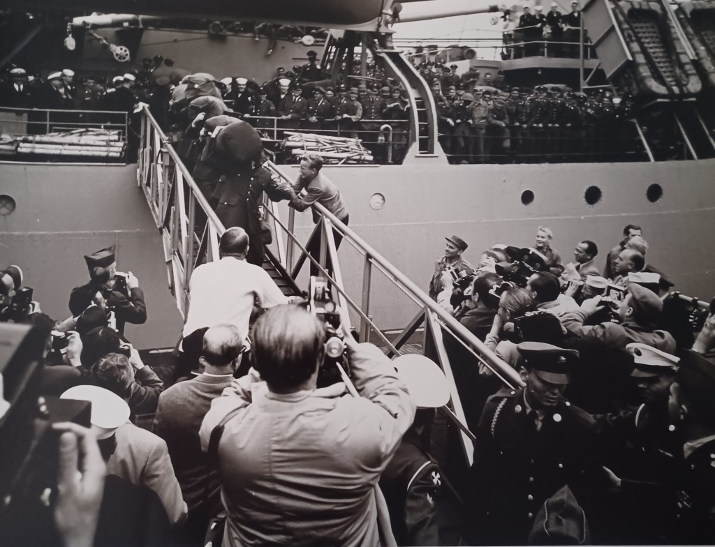 Schwarz-Weiß-Foto von der Ankunft von Elvis Presley in Bremerhaven am 1. Oktober 1958, mit Menschenmengen, die ihn begrüßen, während er die Gangway des Schiffs hinuntersteigt.