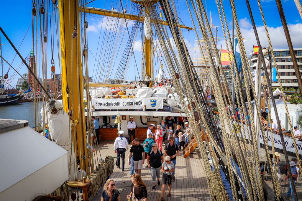 Blick auf das Segelschulschiff Gorch Fock während des Windjammerfestivals, mit Besuchern, die an Deck spazieren. Im Hintergrund sind weitere Schiffe und moderne Gebäude zu sehen.