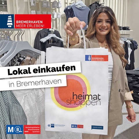 Eine Frau mit langen, braunen Haaren hält eine Einkaufstasche in einem Bekleidungsgeschäft. Im Hintergrund sind Regale mit Kleidung sichtbar. Der Text auf der Tasche sagt 'Lokal einkaufen in Bremerhaven' und 'heimat shoppen'.