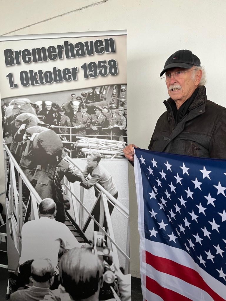 Ein Mann hält eine Fahne der USA und steht neben einem Banner, das an die Ankunft von Elvis Presley in Bremerhaven am 1. Oktober 1958 erinnert.