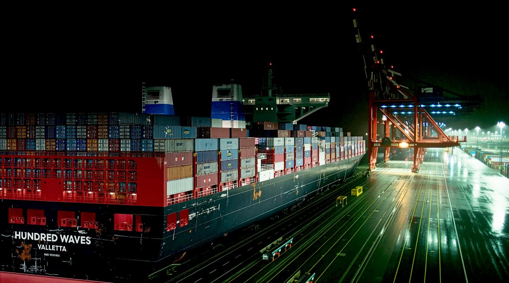 Das Containerschiff "Hundred Waves" im Hafen von Bremerhaven bei Nacht, beladen mit zahlreichen Containern und umgeben von Kranen.