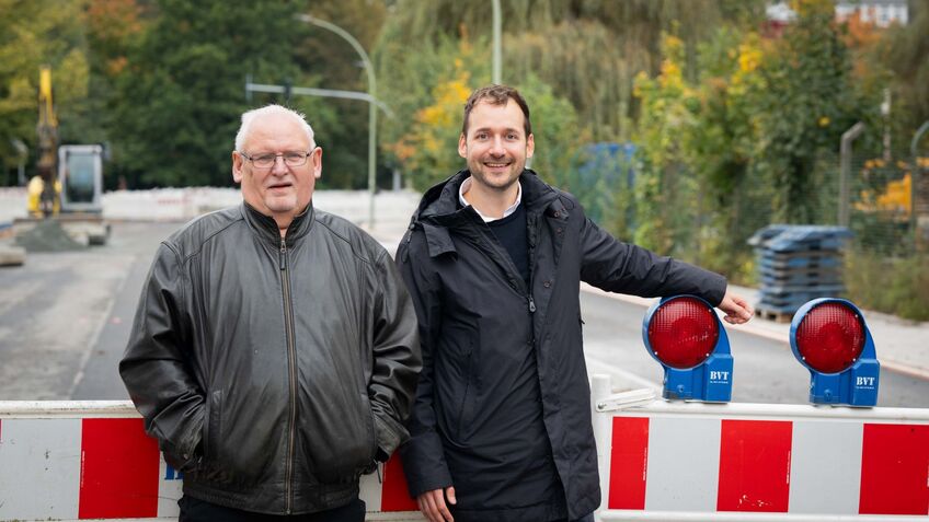 Zwei Männer stehen an einer Baustelle, neben einem Bauzaun mit roten Blinkleuchten. Sie lächeln zufrieden, während im Hintergrund die unvollendete Straße zu sehen ist.