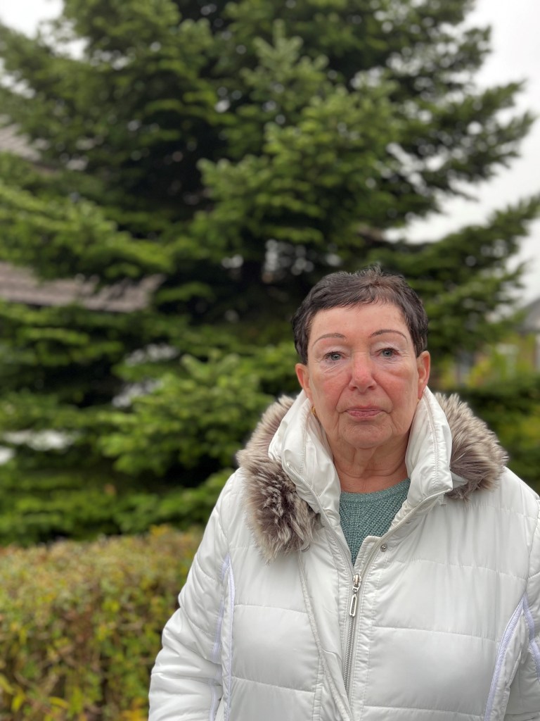 Eine Frau in einem weißen Wintermantel mit einem Fellkragen steht vor einem immergrünen Baum, umgeben von grünen Sträuchern.