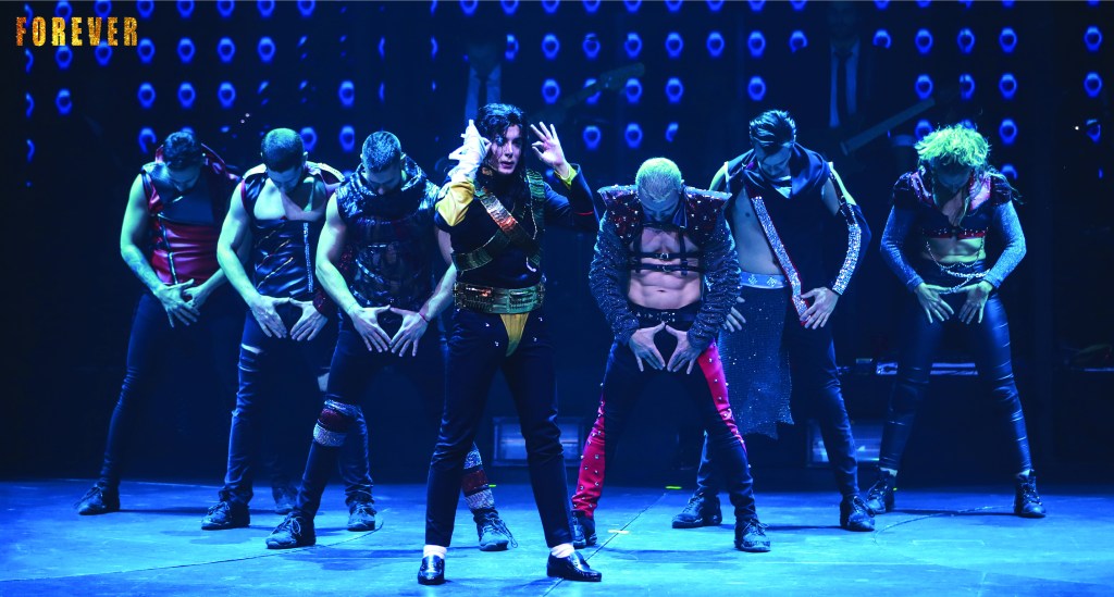 Theatrical performance from 'FOREVER – The Best Show About the King of Pop', featuring a lead dancer surrounded by backup performers, all dressed in stylish, dynamic costumes. The stage is brightly lit with vivid colors, creating an energetic atmosphere.