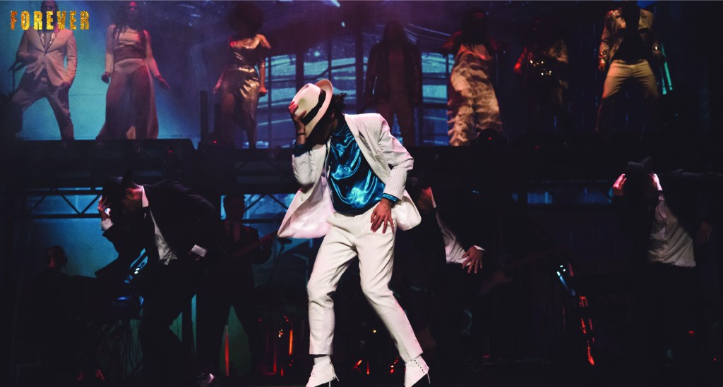 Ein Tänzer in einem weißen Anzug und einem Hut führt eine Choreografie auf einer Bühne auf, umgeben von weiteren Tänzern in einem energiegeladenen Show-Ambiente.