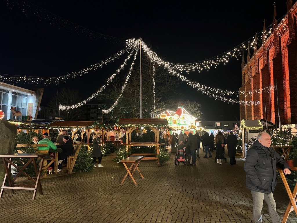A festive night market scene with stalls decorated with Christmas lights, people walking around, and a large building illuminated in the background.