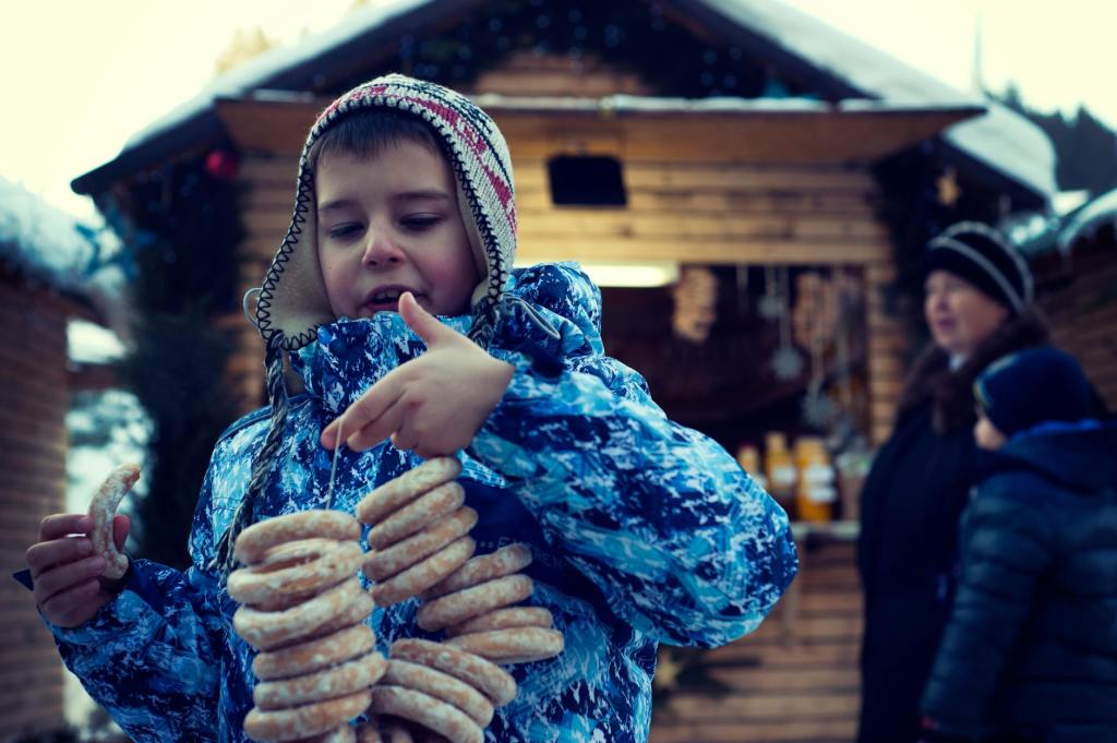 Ein Junge in einem blauen Schneeanzug hält mehrere dekorative Lebkuchenringe in der Hand, während im Hintergrund eine Holzstand mit Weihnachtsangeboten zu sehen ist.