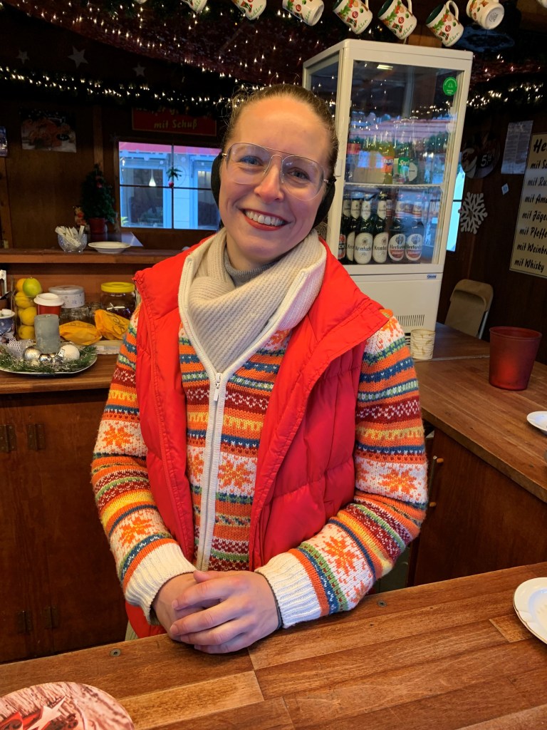 Lächelnde Person in einem Holzstand des Weihnachtsmarktes mit einem rot gefütterten Anorak und buntem Pullover steht hinter einem Tisch, auf dem Getränke und Leckereien sichtbar sind.