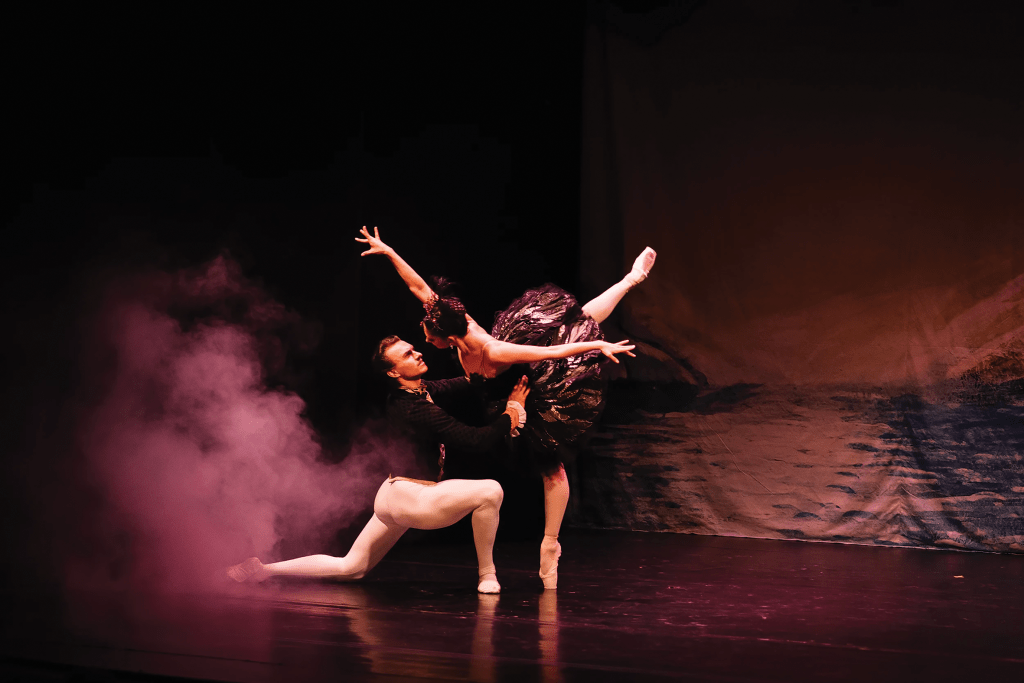 Zwei Tänzer führen eine Ballettaufführung auf einer Bühne mit Nebel durch, einer in einem schwarzen Kostüm und der andere in einem weißen.