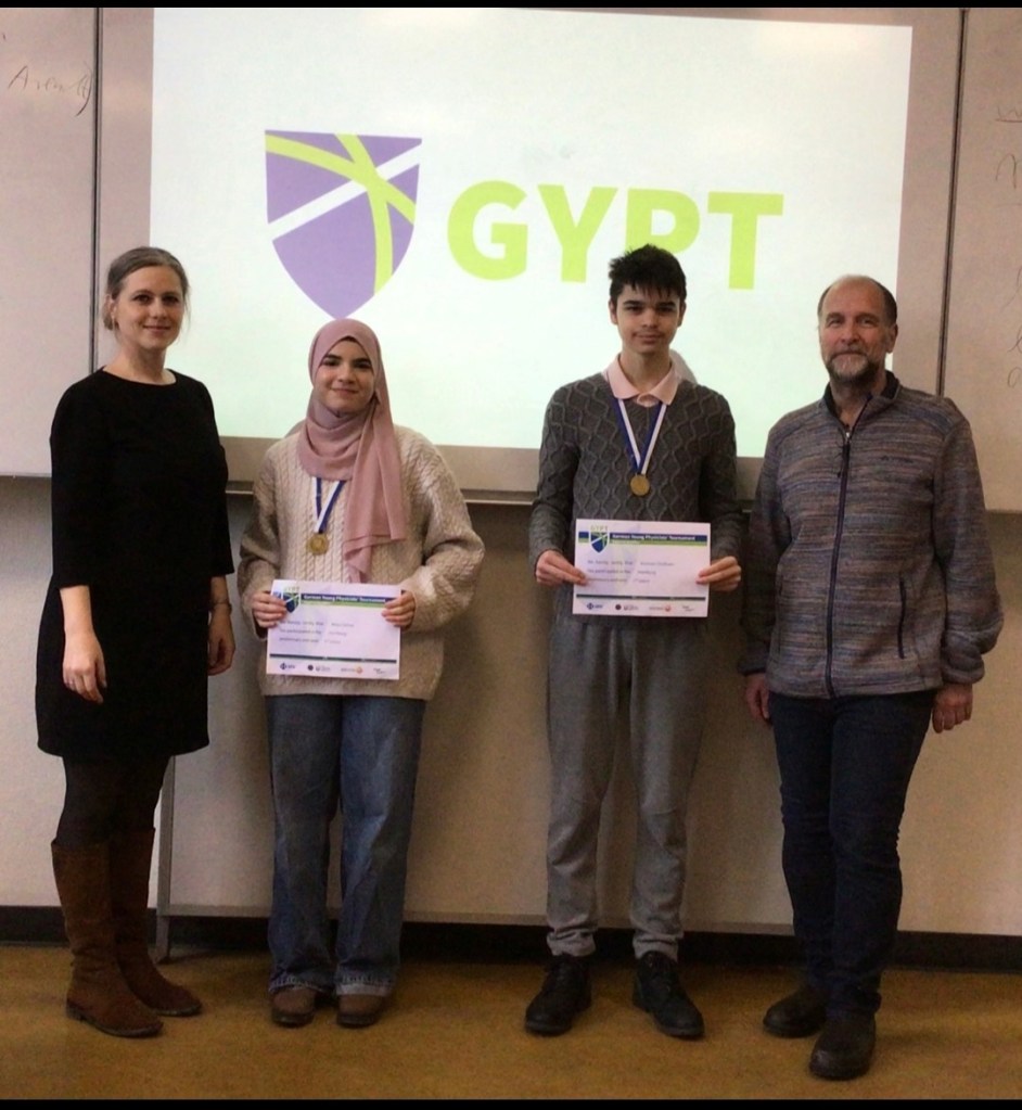 Vier Personen stehen vor einer Leinwand mit dem Logo 'GYPT'. Zwei Schüler halten Urkunden und Medaillen in der Mitte, während zwei Erwachsene an den Seiten stehen.